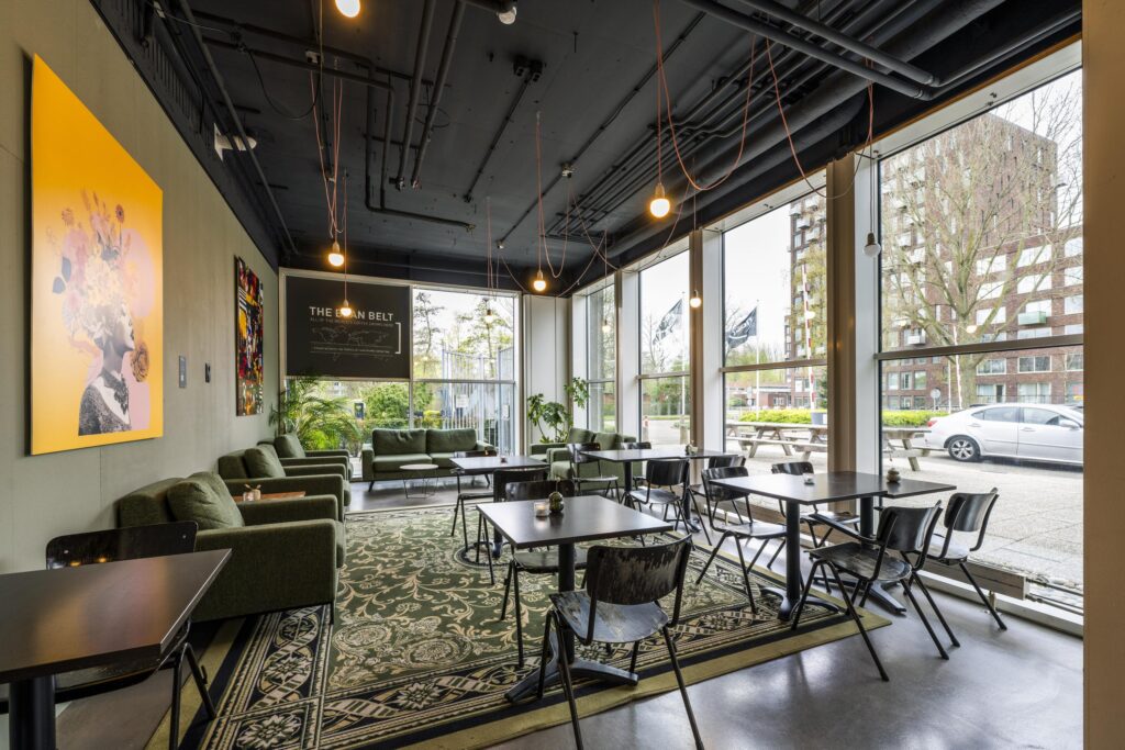 Modern café interior with large windows, dark ceiling, green lounge chairs, and a view of Laan van Kronenburg outside.